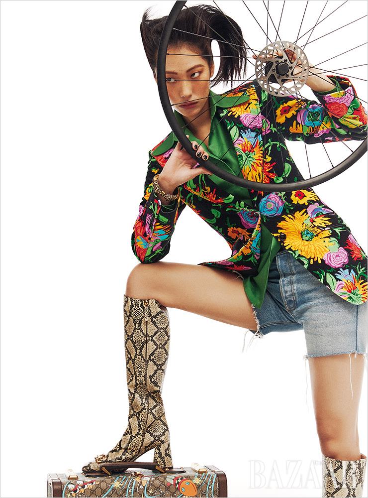 플라워 패턴 재킷, 블라우스, 데님 쇼츠, 팔찌, 파이톤 부츠, 수트케이스는 모두 Gucci.