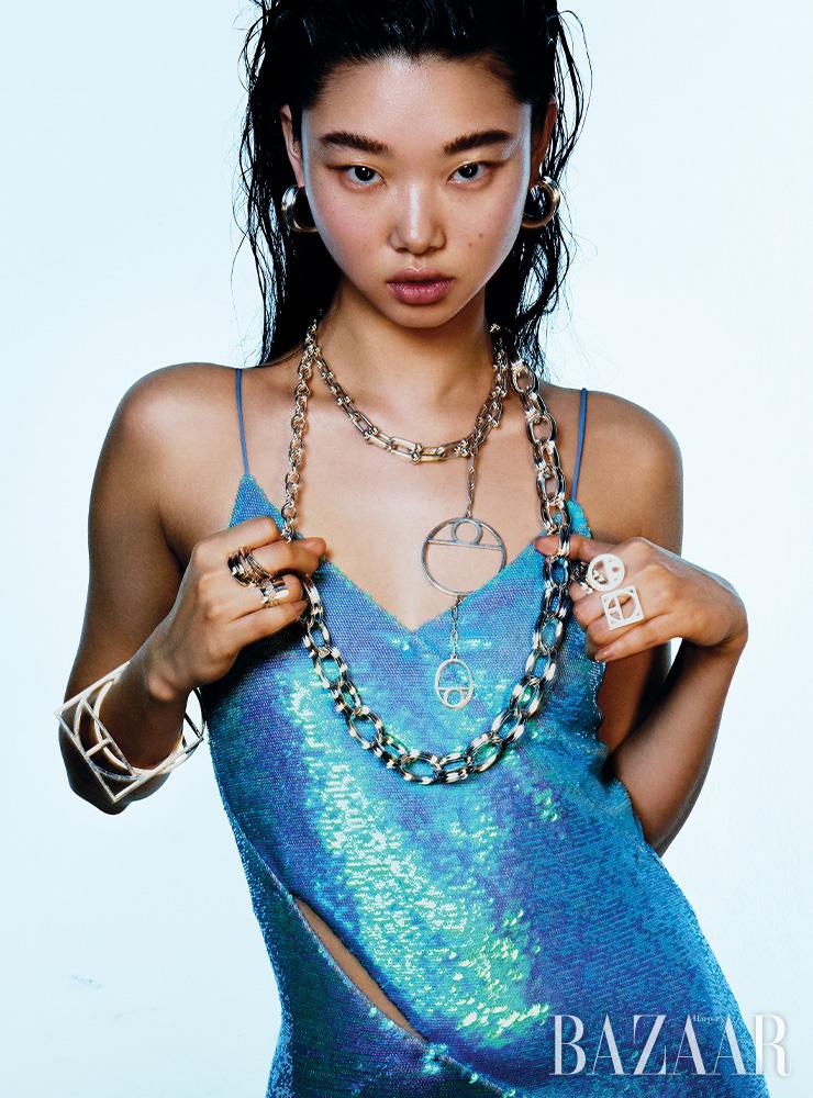 후프 귀고리는 Hermes. 링크 장식의 ‘하드웨어 그레듀에이티드 링크’ 목걸이는 Tiffany & Co.. 링크 목걸이에 연결한 도형 목걸이, 볼드한 체인 목걸이, 오른팔에 착용한 팔찌, 왼손에 낀 도형 반지는 모두 Hermes. 왼손에 함께 낀 볼드한 십자가 반지는 Chrome Hearts. 오른손 중지에 낀 반지들은 세트로 29만8천원 Arsenix. 약지에 낀 글리터 디테일 반지는 28만원 Jem & Pebbles.컷아웃 디테일 드레스는 1백65만원 Off-White.